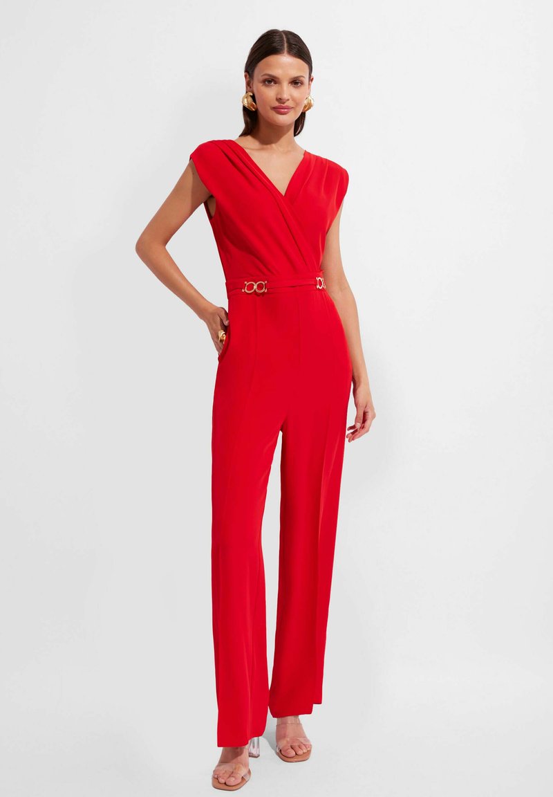 Czerwony kombinezon z dekoltem w serek, rękawami na ramionach i akcentującym złotym paskiem; dopasowana góra i szerokie nogawki; gładka faktura materiału.