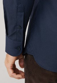 Man in een marineblauw shirt met kleine witte stippen en een bruine broek, die de details van de mouwen en zijnaad toont.