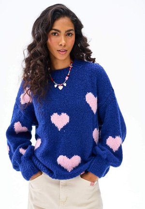 Kobieta o długich kręconych włosach, ubrana w niebieski sweter w różowe serduszka oraz beżowe spodnie, ze złożonymi rękami w kieszeniach, na białym tle.