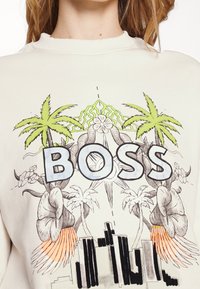 Krémová mikina s grafikou, která zobrazuje palmy, květiny a slovo "BOSS" v modré barvě, doplněná o detaily v zelené a oranžové.