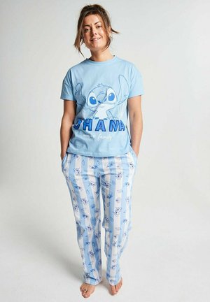 Frau mit hochgestecktem Haar trägt ein blaues T-Shirt mit Stitch-Motiv und passende hellblaue und weiße gestreifte Schlafanzughose, steht barfuß mit den Händen in den Taschen.