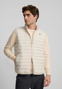 Chaleco acolchado beige con cuello alto y cremallera frontal, con costuras horizontales y un emblema en el pecho. Lleva puesto sobre un suéter ligero.