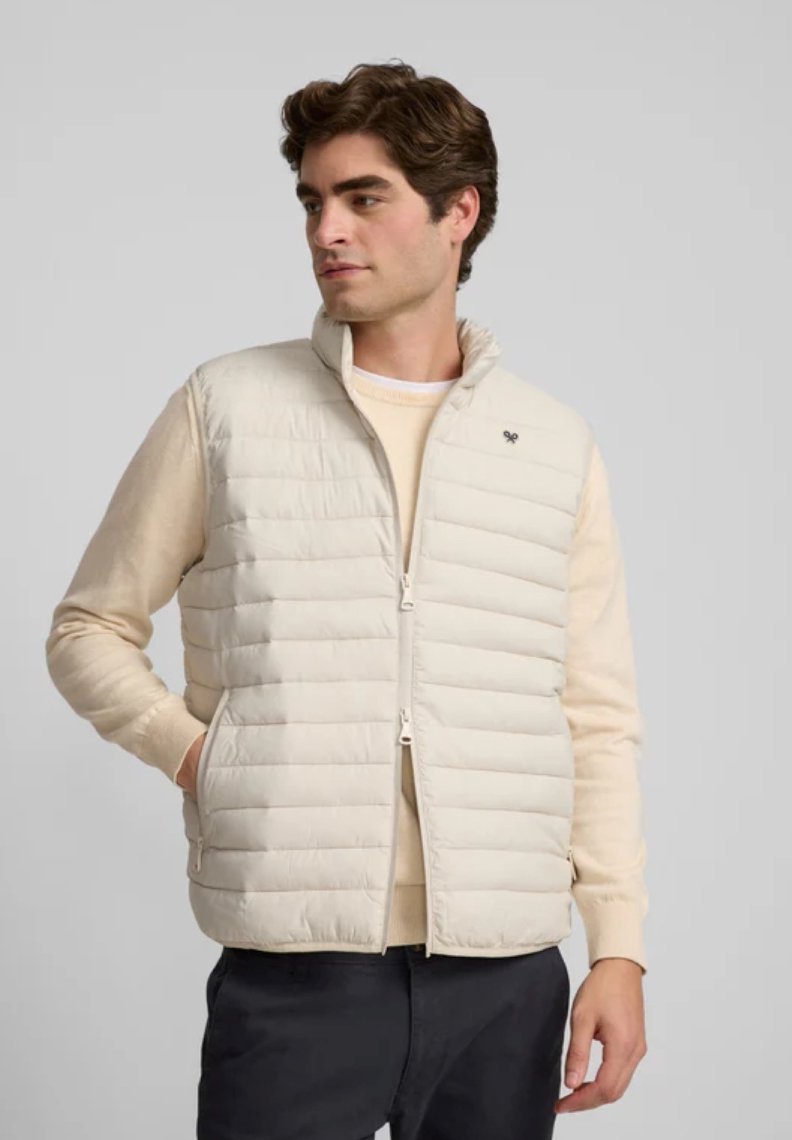 Chaleco acolchado beige con cuello alto y cremallera frontal, con costuras horizontales y un emblema en el pecho. Lleva puesto sobre un suéter ligero.