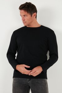 Juodas ilgesnis marškinėliai su apvalia iškirpte, pagaminti iš lygaus audinio. Modelis vilki pilkas džinsus ir stovi priešais vientisą fono.