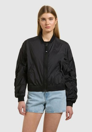 Fiatal nő hosszú szőke hajjal, fekete cipzáros bomber dzsekit és világoskék farmer rövidnadrágot viselve, egyszínű háttér előtt.