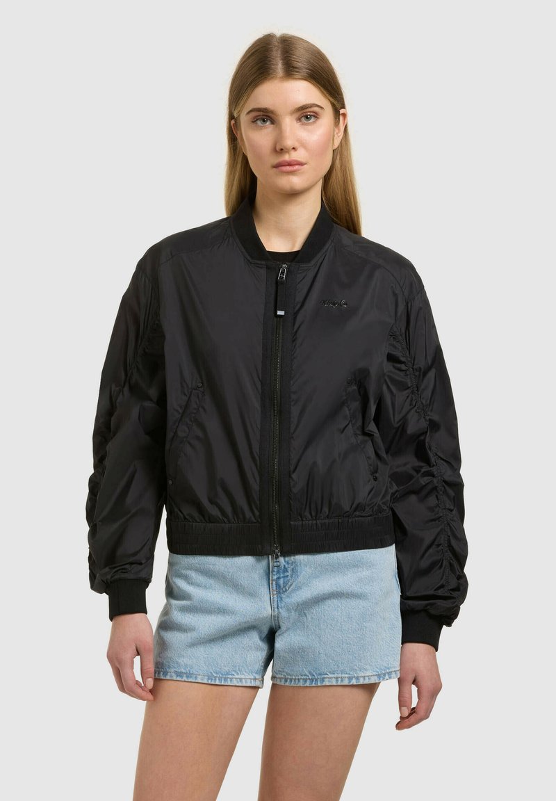 Mlada ženska z dolgimi blond lasmi, oblečena v črno bomber jakno z zadrgo in svetlo modre denim kratke hlače, na enobarvnem ozadju.