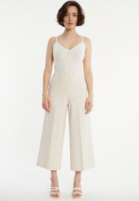 Weißes Trägertop kombiniert mit hoch geschnittenen, weit geschnittenen beigen Hosen mit vertikalen Nadelstreifen. Das Model trägt geschlossene weiße Sandalen.