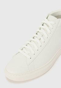 Zapatilla de cuero blanca con un acabado texturizado, que presenta costuras laterales prominentes, cordones planos y una suela de goma. Logo de la marca visible en el costado.