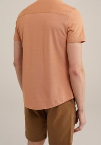 Tričko s krátkým rukávem a strukturovaným výstřihem do kulata v světle oranžové barvě, s jemným vzorem, doplněné hnědými šortkami. Hladká tkanina a volný střih.