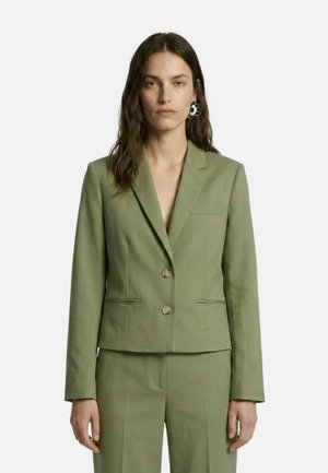 Vrouw met lang donker haar draagt een lichtgroene op maat gemaakte blazer en bijpassende broek, staand voor een effen lichte achtergrond.
