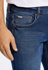 La mano sinistra di un uomo riposa accanto alla tasca frontale di un paio di jeans blu scuro con etichetta del marchio visibile, indossando una camicia bianca.