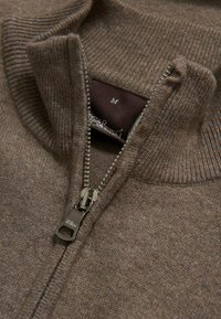 Maglione zip up marrone con colletto a coste, dotato di una cerniera metallica e di un'etichetta con la taglia. La texture è morbida con una finitura lavorata a maglia.