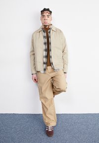 Beige jakke over en brun sweater, lysebrune cargo bukser og brune sneakers med hvide snørebånd. Model står imod en ensfarvet hvid væg.