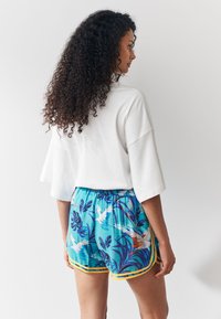 Maglietta bianca oversize abbinata a pantaloncini blu con stampa tropicale colorata e rifinitura gialla, caratterizzati da una vestibilità comoda e vita elastica.
