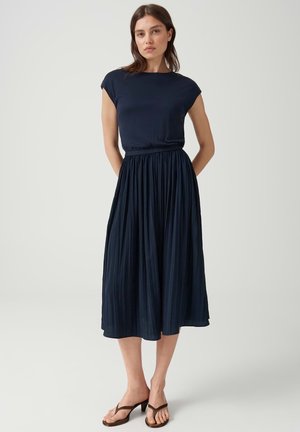 Femme portant un haut bleu marine à manches courtes et une jupe midi plissée, debout les mains dans le dos sur un fond uni.