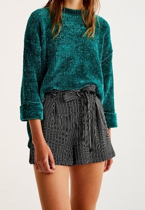 Kvinde iført en grøn tekstureret langærmet sweater og højtaljede sorte og hvide shorts med lodrette striber og bundet bælte.
