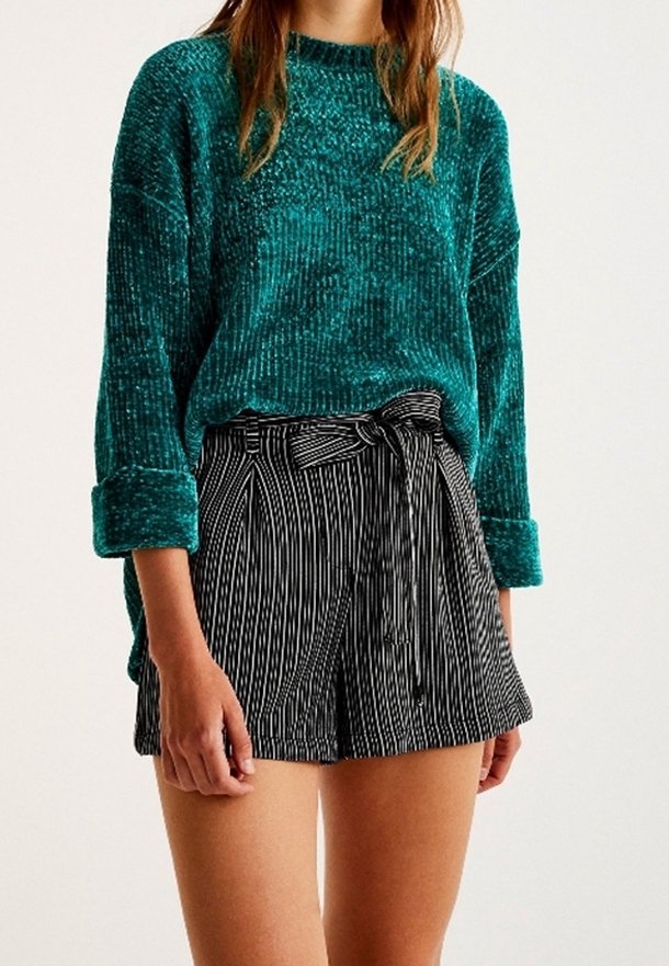 Femme portant un pull vert texturé à manches longues et un short taille haute rayé vertical noir et blanc avec une ceinture nouée.