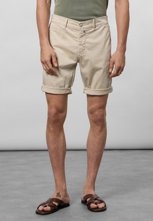 Beige katoenen shorts met opgerolde boorden, voor knoopsluiting en zijzakken. Gepaard met donkerbruine sandalen. Effen achtergrond.