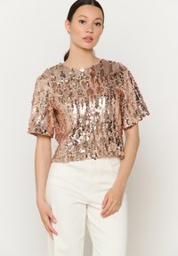Sequin top in rose gold with short sleeves, round neckline, and a cropped design. Worn with white high-waist trousers.