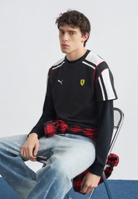 Νεαρός με κατσαρά μαλλιά κάθεται σε καρέκλα φορώντας μαύρο μπλουζάκι Puma Ferrari πάνω από μακριά μαύρα μανίκια, ανοιχτόχρωμο τζιν και κόκκινο καρό πουκάμισο δεμένο στη μέση.