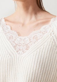 Pletený svetr v krémové barvě s výstřihem do V, zdobený květinovou krajkou u výstřihu. Materiál má strukturovaný žebrovaný vzor.