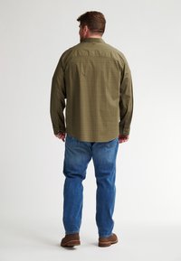 Camicia verde oliva a maniche lunghe con un motivo sottile, polsini con bottone e orlo dritto, abbinata a jeans blu e stivali marroni.