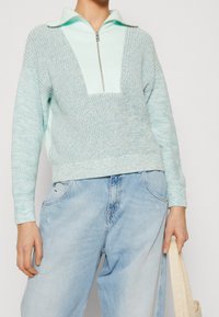 Hellblauer Strickpullover mit V-Ausschnitt und Reißverschluss-Kragen, aus strukturiertem Stoff. Kombiniert mit hellblauen, locker sitzenden Jeans.