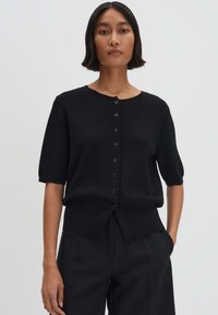 Black knitted short-sleeve cardigan with a scalloped neckline, featuring a row of black buttons down the front and a relaxed fit.