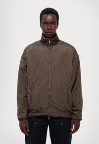 Giacca in nylon marrone con colletto alto, zip frontale e due tasche laterali. Presenta una trama liscia e un branding minimale.