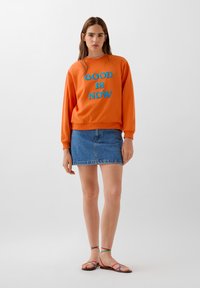 Felpa arancione con testo blu "GOOD IS NOW", abbinata a una mini gonna in denim e sandali multicolori con lacci. Tessuto morbido, vestibilità rilassata.