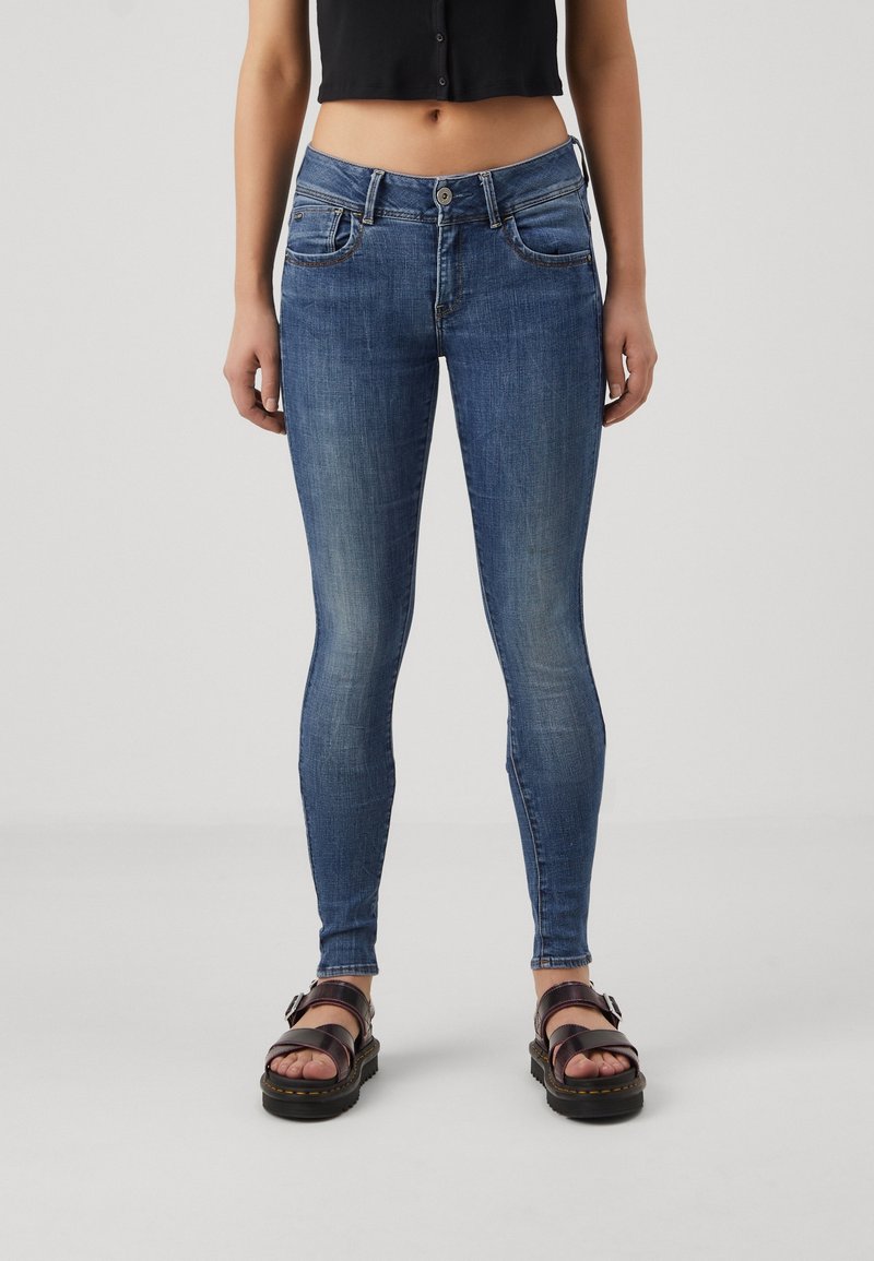 Wąskie dżinsy w średnim niebieskim kolorze denimu, o dopasowanym kroju, z pięcioma kieszeniami i subtelnymi efektami przetarć. Zestawione z ciemnymi sandałami.