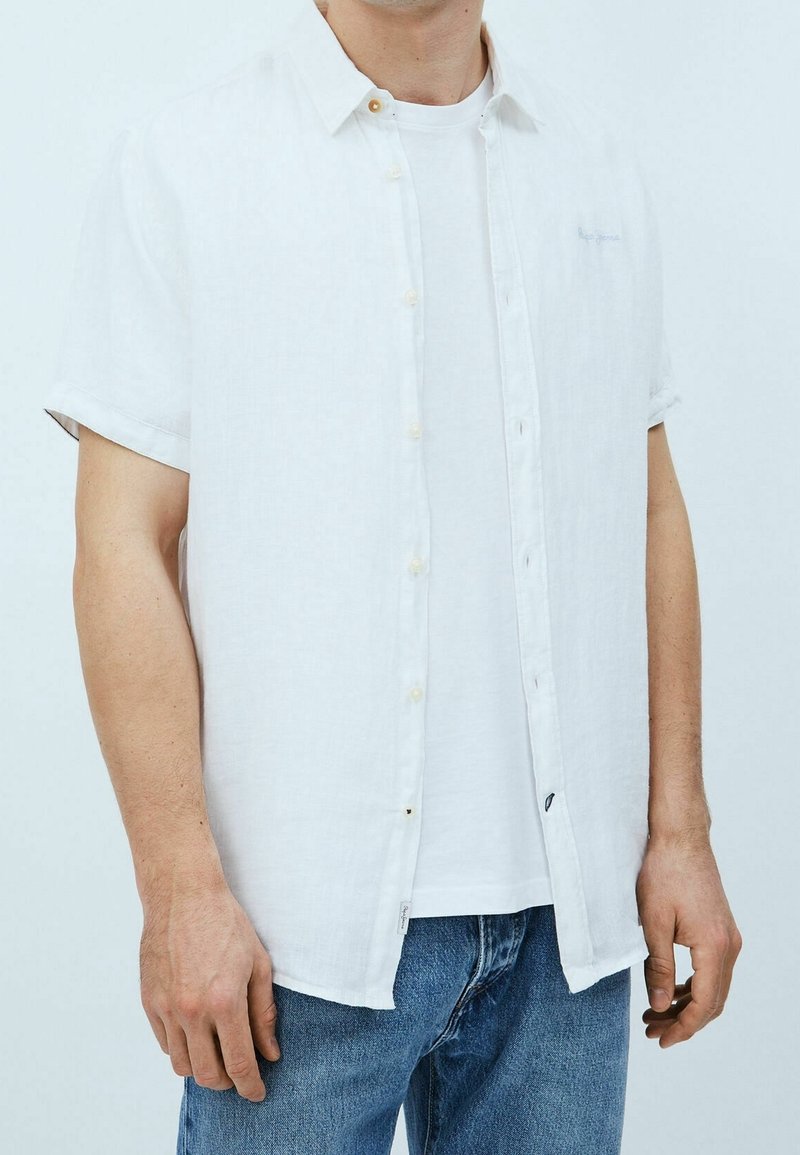 Homme portant une chemise blanche à manches courtes boutonnée par-dessus un t-shirt blanc avec un jean bleu, debout devant un fond uni.