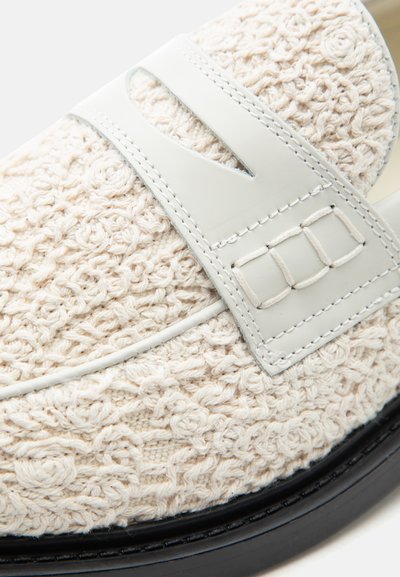 Mocassin en maille texturée couleur crème avec une bride en cuir blanc et une semelle noire, montrant des coutures détaillées et des motifs tissés.