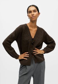 Cardigan en tricot marron avec un col en V, fermeture à nouer et motif texturé. Les manches longues et une coupe décontractée complètent le design.