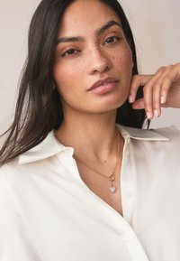 Collier en or avec un pendentif en forme de cœur orné de petites pierres scintillantes. Le mannequin porte une chemise blanche à col ; cheveux longs et foncés.