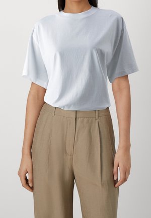 Person trägt ein schlichtes weißes kurzärmeliges T-Shirt, das in beige, hoch taillierte Hosen mit Seitentaschen gesteckt ist.