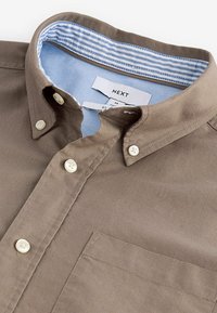 Camisa marrón de botones con un bolsillo en el pecho, que presenta un forro de cuello azul claro con rayas blancas y botones blancos. Diseño limpio y estructurado.