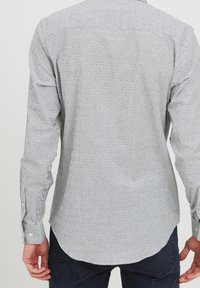 Camicia button-down di un grigio chiaro con sottili righe verticali; il tessuto ha una superficie liscia. Presenta polsini con bottoni e orlo curvo.