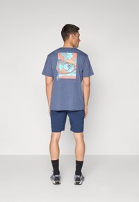 Men's short-sleeve blue t-shirt with a colorful fish graphic on the back, paired with dark blue shorts and athletic shoes.