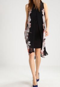 Černé šaty s průhlednou květinovou vrstvou zdobenou bílými a růžovými květy. Design je bez rukávů a má volný tvar. Doplňují je modré podpatky.