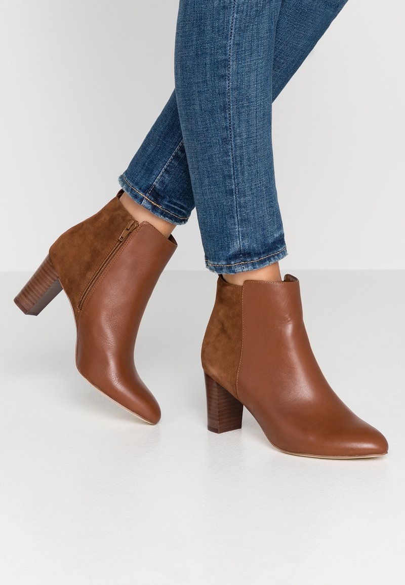 Bottines en cuir et suède marron avec talons carrés, portées avec un jean bleu retroussé sur un sol blanc.