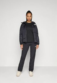 Svart polstret jakke med hette, kombinert med en svart genser med rund hals og svarte bukser. Har logoaksenter, brukt med lyse sko.