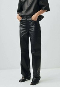 Persona che indossa pantaloni dritti in ecopelle nera e una maglia in ecopelle coordinata, con le mani nelle tasche, in piedi su un pavimento bianco.