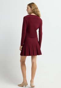 Top de manga larga de canalé color marrón rojizo combinado con una falda plisada del mismo color. La textura es suave, el diseño es ajustado y unos zapatos beige completan el conjunto.