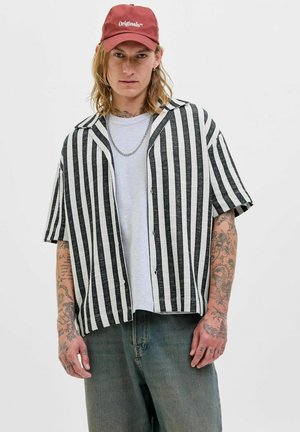Camisa de rayas negra y blanca de manga corta con un corte relajado, con botones en la parte delantera, combinada con una camiseta gris claro y jeans azules desgastados.