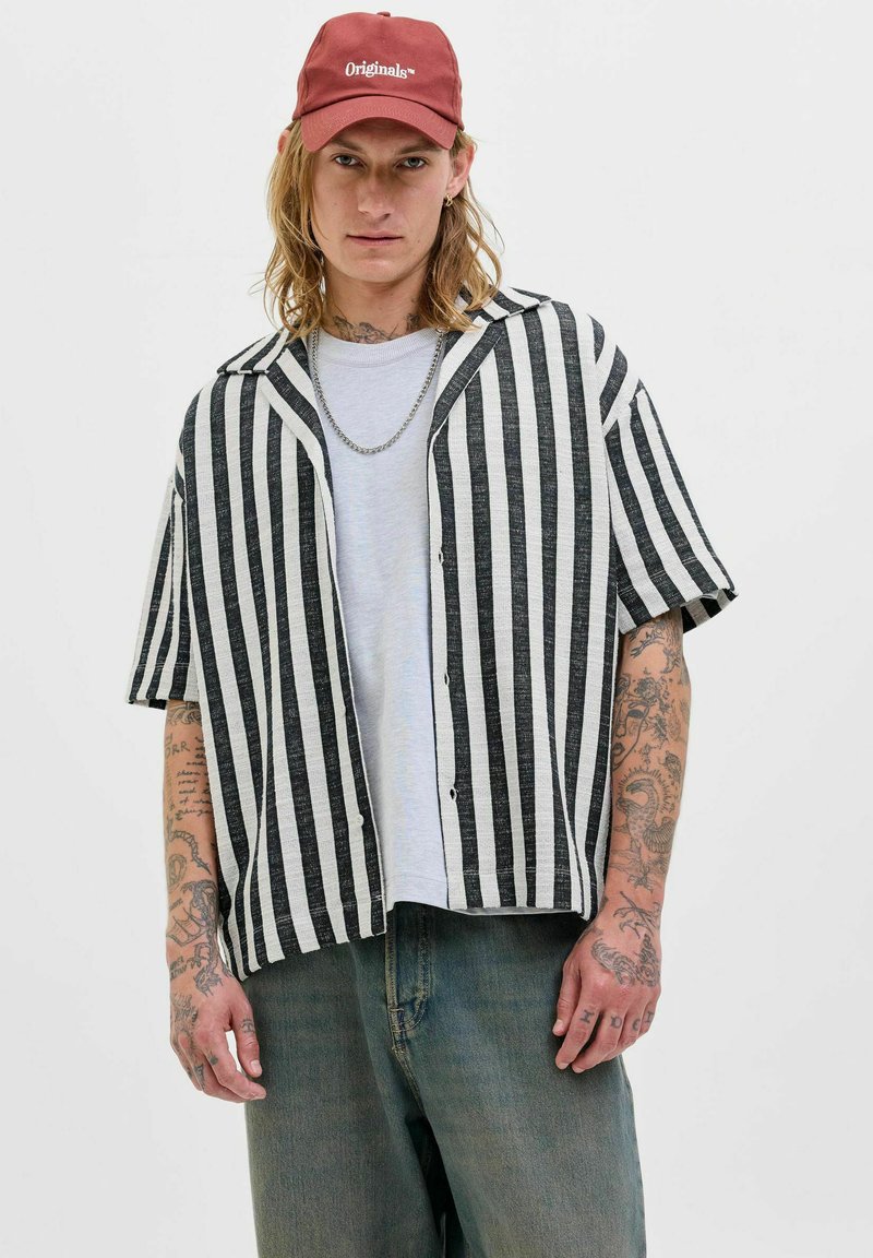 Chemise à rayures noir et blanc à manches courtes, coupe ample, avec des boutons sur le devant, associée à un t-shirt gris clair et à un jean bleu déchiré.