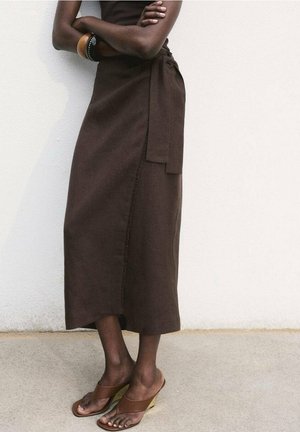 Personne bras croisés portant une robe marron sans manches et des sandales marron à talons hauts, debout contre un mur blanc.