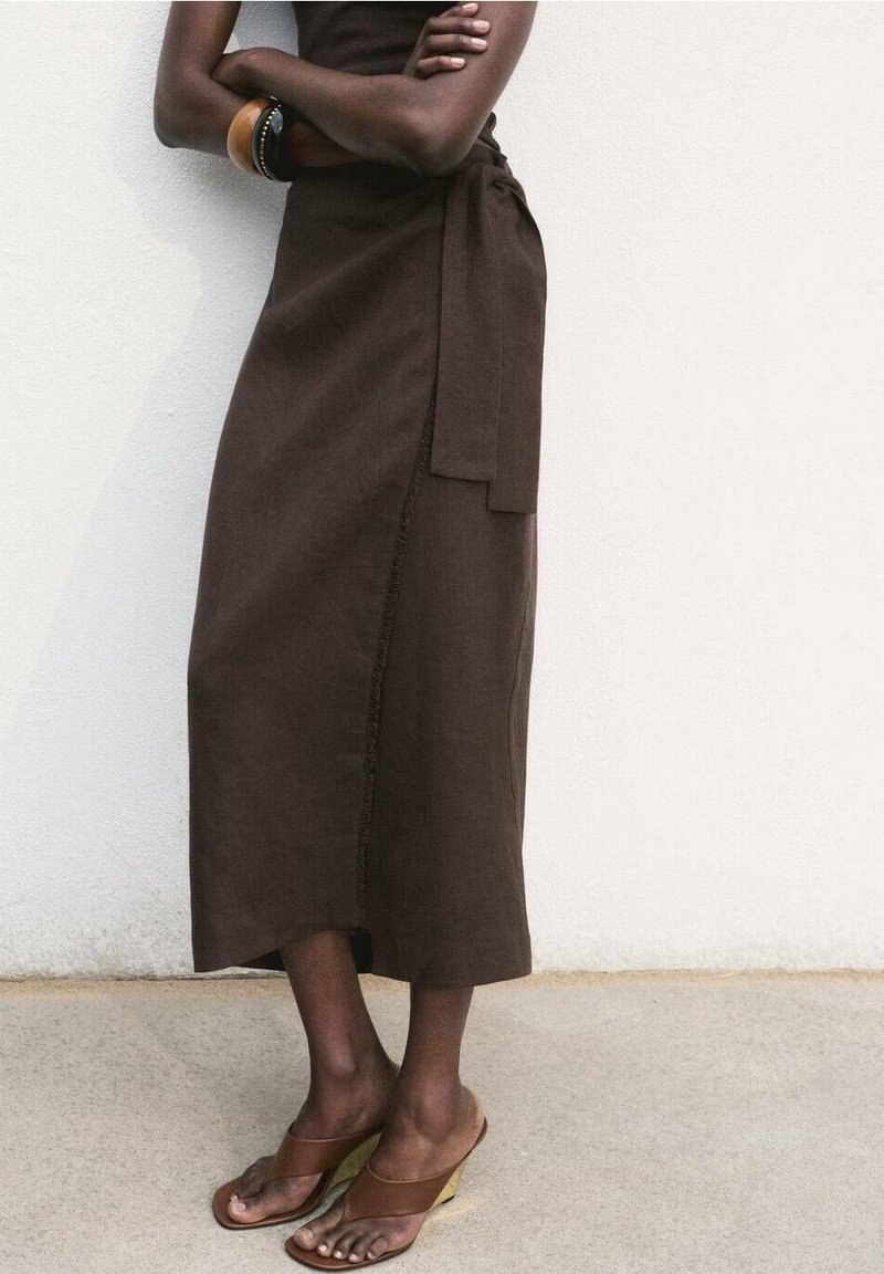Personne bras croisés portant une robe marron sans manches et des sandales marron à talons hauts, debout contre un mur blanc.