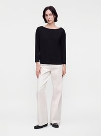 Jeune personne aux cheveux courts et foncés, portant un haut noir à manches longues, un pantalon blanc à jambes larges et des chaussures noires, debout devant un fond blanc uni.