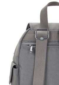 Sac à dos gris avec une sangle tissée et une boucle en métal, comportant une poche zippée et une poche à rabat sur le côté sur fond blanc.